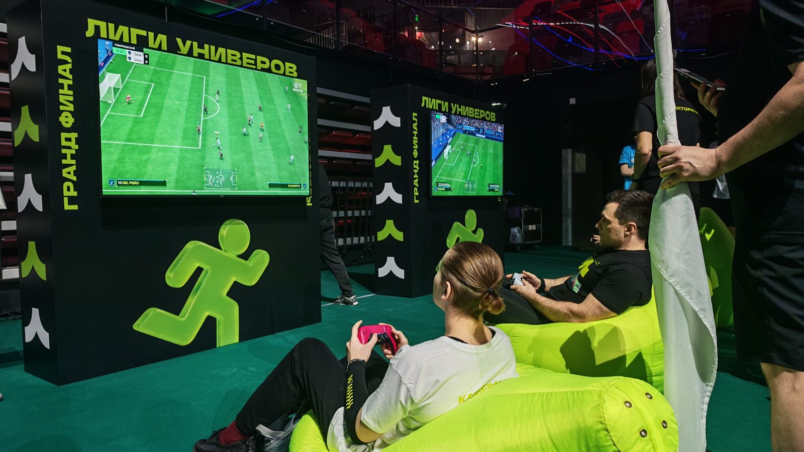 Зона футбола на PlayStation 5: двое участников с геймпадами перед двумя телевизорами в брендинге гранд-финала «Лиги Универов»