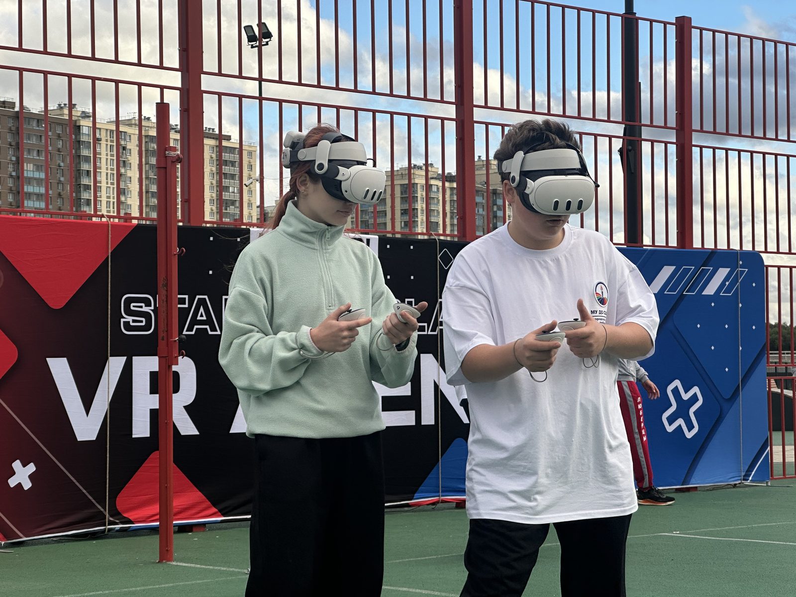 Два участника в VR-шлемах с контроллерами крупным планом на фоне баннера арены