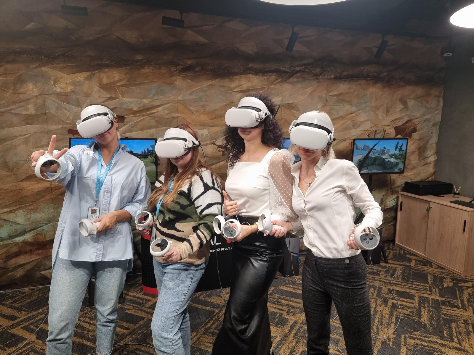 Четыре сотрудницы в VR-шлемах на корпоративе, позади экраны с трансляцией