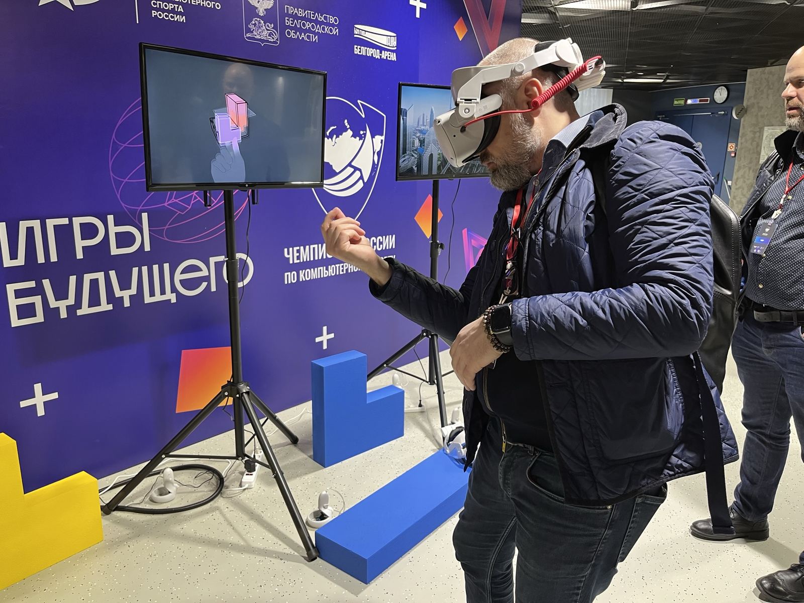 Гость выставки играет в VR, рядом два экрана с трансляцией на треногах