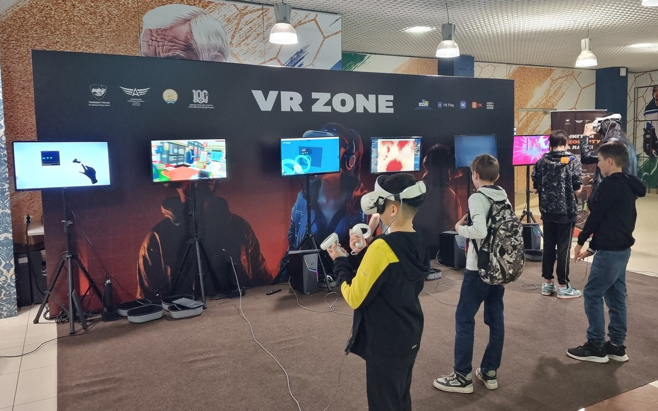 Большая брендированная VR-зона на площадке: ряд экранов с трансляцией вдоль стенда, игроки в шлемах и зрители